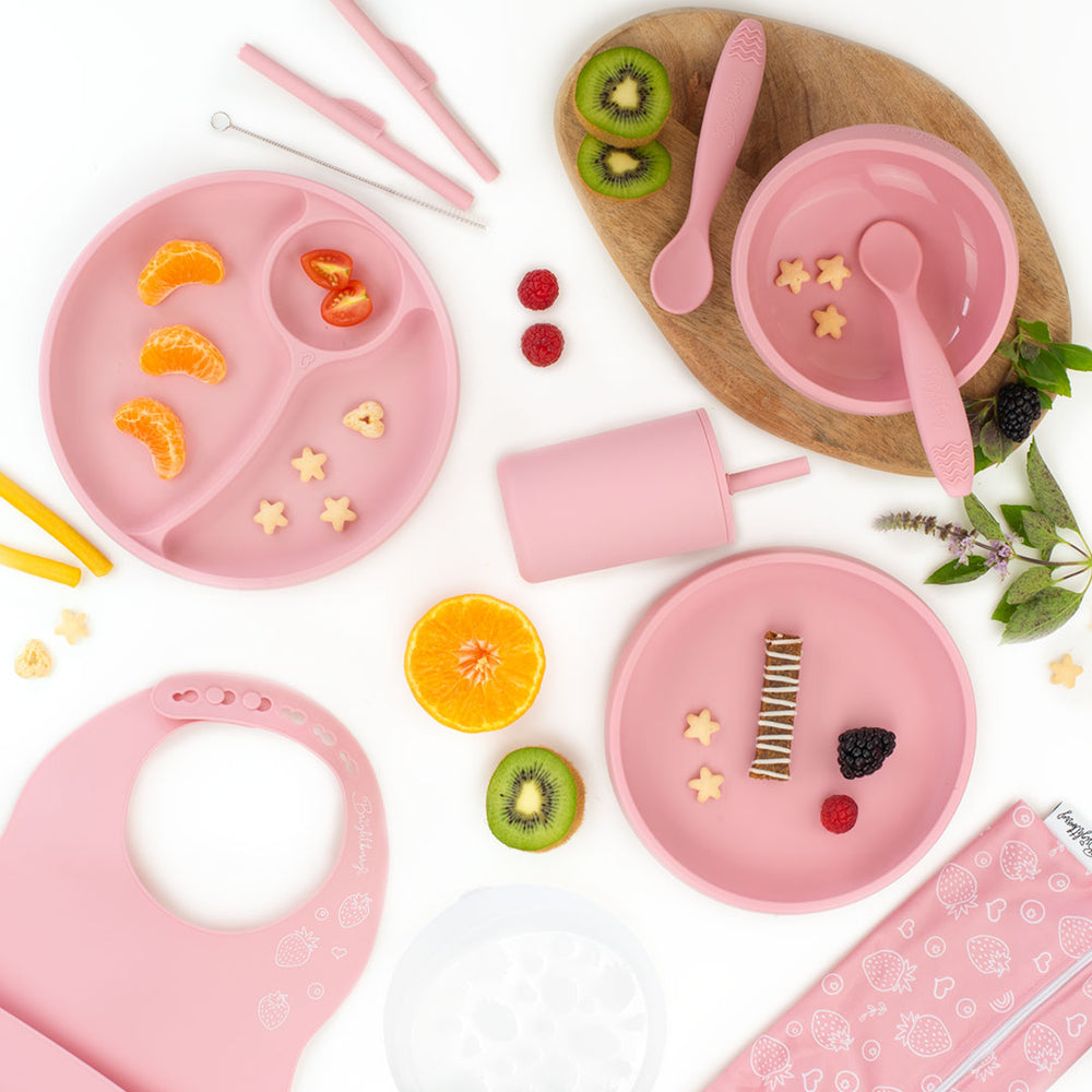 Flat lay image of a coral pink silicone feeding set for kids, including divided plates, bowls, spoons, a smoothie cup with a lid, a bib, and a straw cleaning brush, surrounded by fruit slices and snacks, with a light wooden board underneath and a white background.