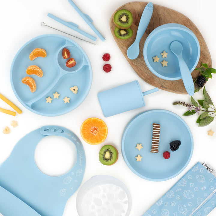 Flat lay image of a pacific blue silicone feeding set for kids, including divided plates, bowls, spoons, a smoothie cup with a lid, a bib, and a straw cleaning brush, surrounded by fruit slices and snacks, with a light wooden board underneath and a white background.