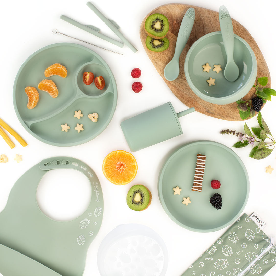 Flat lay image of a green silicone feeding set for kids, including divided plates, bowls, spoons, a smoothie cup with a lid, a bib, and a straw cleaning brush, surrounded by fruit slices and snacks, with a light wooden board underneath and a white background.