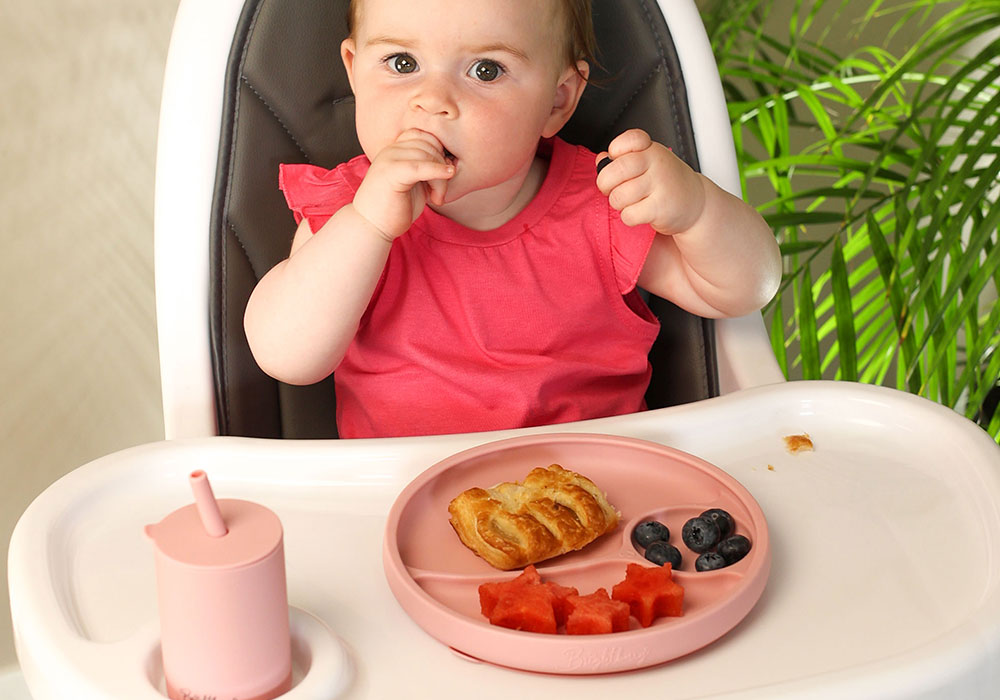 baby girl self feeding from silicone suction plate with dividers, and smoothie cup for kids, sitting in a high chair